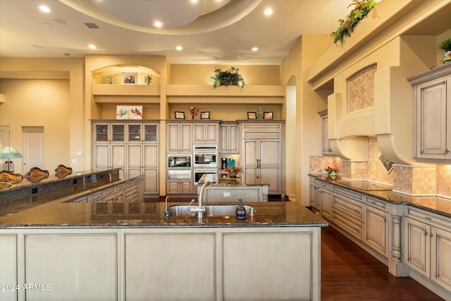 kitchen with black electric stovetop, dark stone counters, built in shelves, a spacious island, and sink