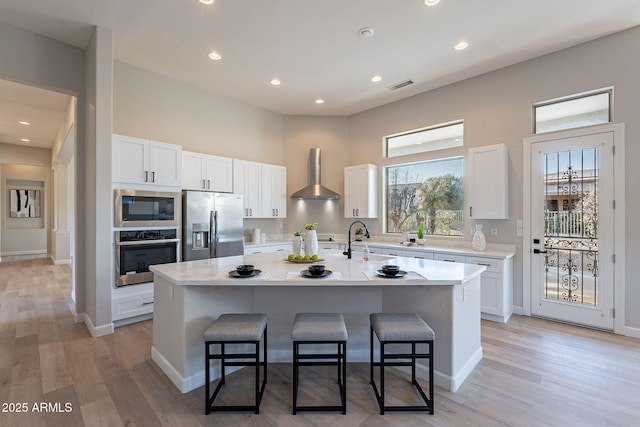 kitchen with wall chimney range hood, a kitchen breakfast bar, an island with sink, white cabinets, and appliances with stainless steel finishes