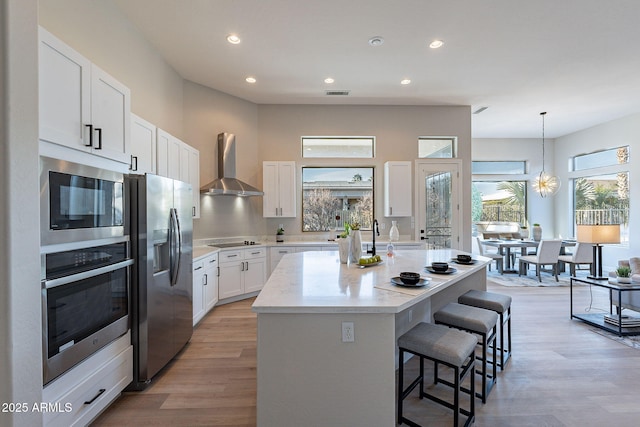 kitchen with appliances with stainless steel finishes, wall chimney range hood, light hardwood / wood-style flooring, white cabinetry, and an island with sink