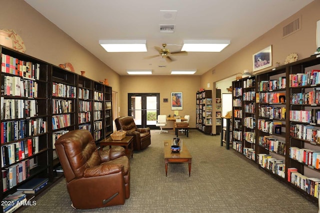 living area with wall of books, carpet, visible vents, and ceiling fan