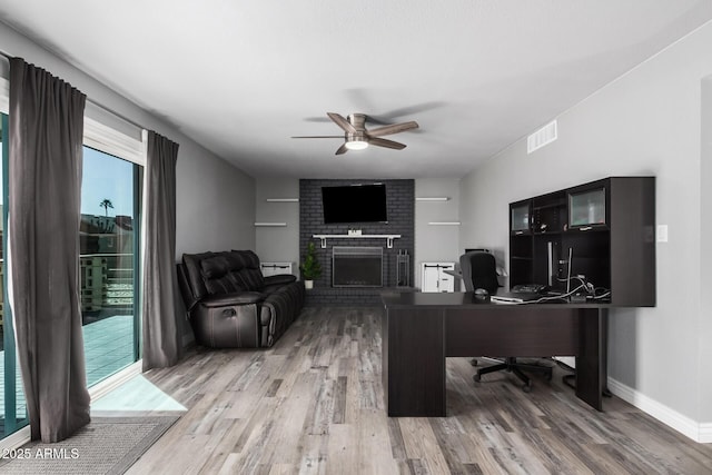 office with visible vents, a brick fireplace, baseboards, light wood-style flooring, and a ceiling fan