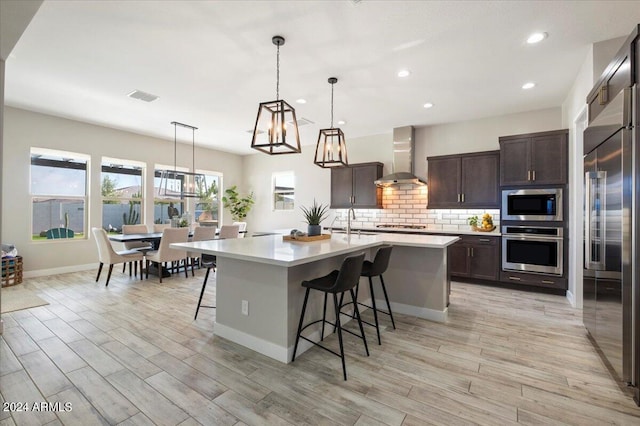 kitchen with wall chimney exhaust hood, stainless steel appliances, hanging light fixtures, a kitchen island with sink, and a breakfast bar area