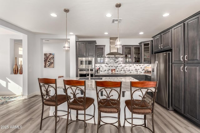 kitchen featuring stainless steel appliances, hanging light fixtures, wall chimney range hood, and a center island with sink