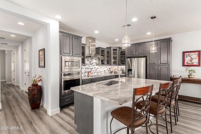 kitchen featuring sink, decorative light fixtures, appliances with stainless steel finishes, an island with sink, and wall chimney range hood