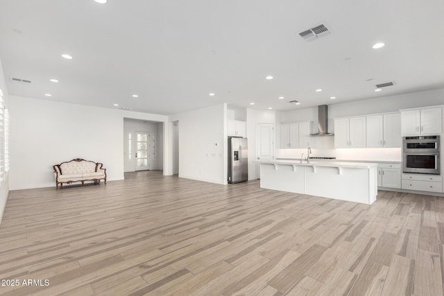 kitchen featuring wall chimney range hood, a kitchen island with sink, stainless steel appliances, white cabinets, and light wood-type flooring