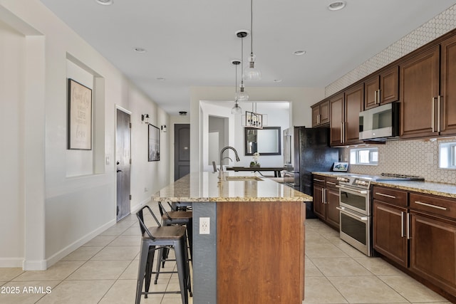 kitchen featuring light tile patterned floors, a center island with sink, a sink, stainless steel appliances, and tasteful backsplash