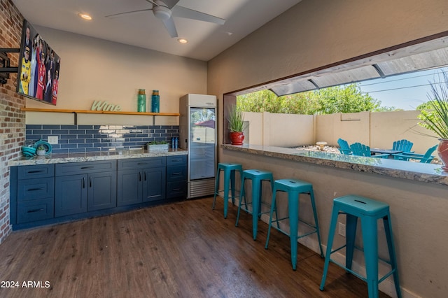 kitchen with ceiling fan, light stone counters, dark hardwood / wood-style floors, decorative backsplash, and a breakfast bar