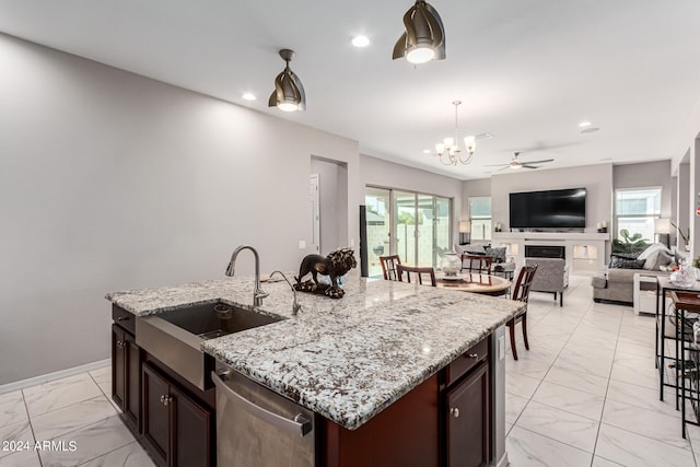 kitchen with light stone counters, ceiling fan with notable chandelier, dishwasher, a center island with sink, and sink