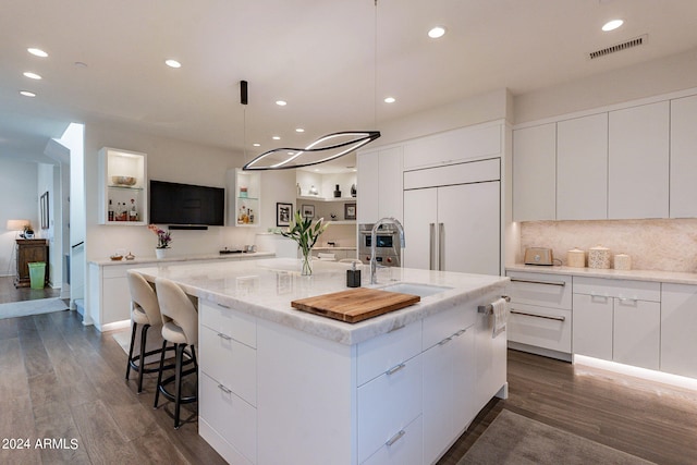 kitchen with sink, dark hardwood / wood-style floors, paneled built in fridge, a kitchen island with sink, and white cabinets