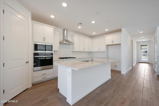 kitchen with sink, white cabinets, a kitchen island with sink, stainless steel appliances, and wall chimney exhaust hood