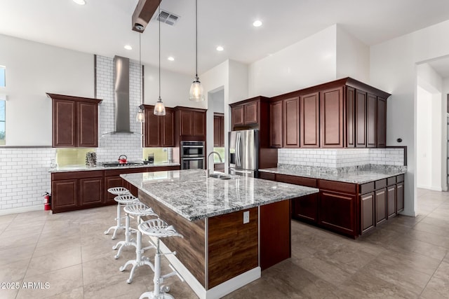 kitchen with hanging light fixtures, an island with sink, stainless steel appliances, wall chimney exhaust hood, and light stone counters