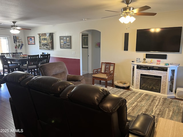 living area with ceiling fan, arched walkways, a stone fireplace, and wood finished floors