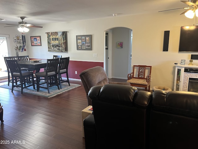 living room with dark wood finished floors, arched walkways, a stone fireplace, baseboards, and ceiling fan