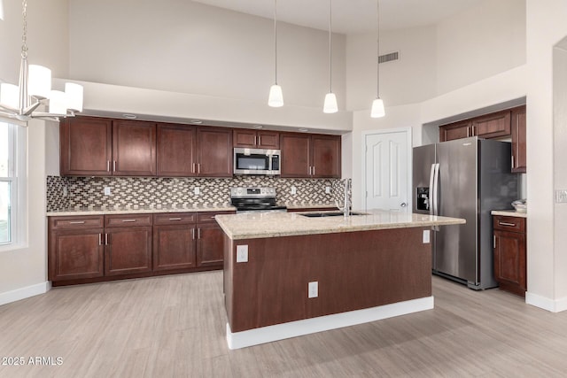 kitchen featuring appliances with stainless steel finishes, a towering ceiling, light stone counters, pendant lighting, and an island with sink