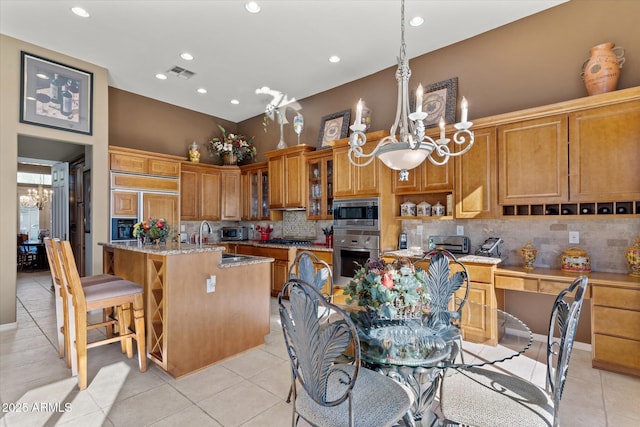 kitchen with sink, an inviting chandelier, light stone counters, built in appliances, and a kitchen island with sink