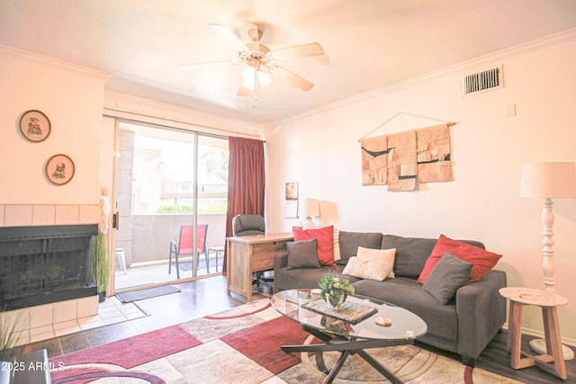 living area with ornamental molding, a ceiling fan, visible vents, a tile fireplace, and wood finished floors