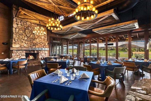 dining area featuring a high ceiling, a stone fireplace, ceiling fan with notable chandelier, and beam ceiling