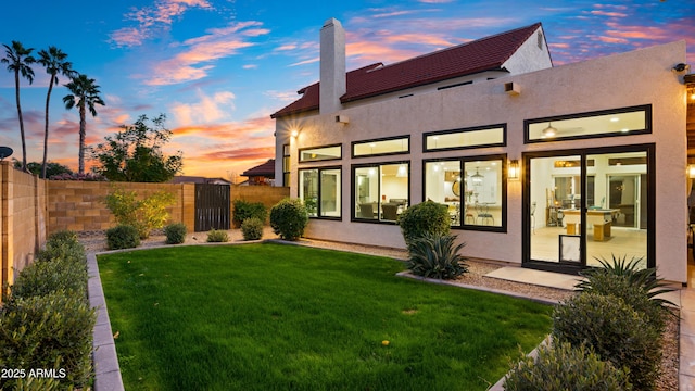 back of house at dusk with a lawn, fence, and stucco siding