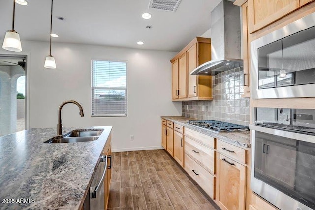 kitchen featuring appliances with stainless steel finishes, light brown cabinets, a sink, wall chimney range hood, and light wood-type flooring