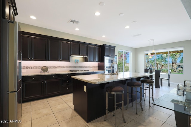 kitchen featuring appliances with stainless steel finishes, decorative light fixtures, an island with sink, sink, and light stone counters