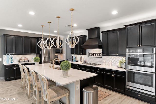 kitchen with dark cabinets, stainless steel appliances, light wood-type flooring, an island with sink, and custom range hood