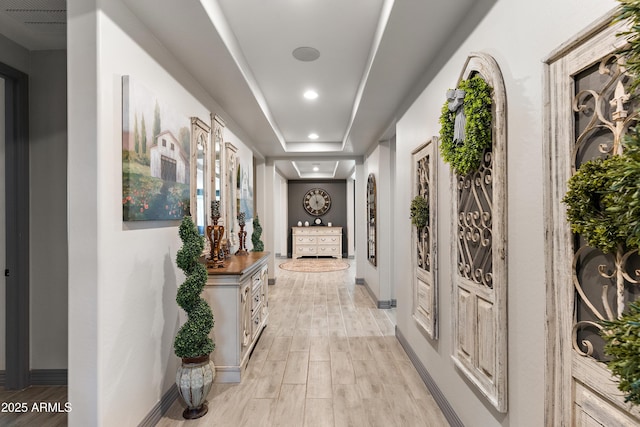 hallway featuring recessed lighting, a raised ceiling, baseboards, and light wood finished floors