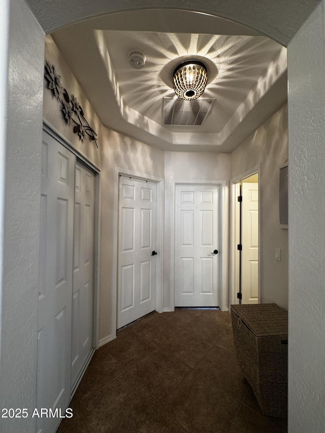 hallway with a tray ceiling, dark tile patterned floors, arched walkways, and a textured wall