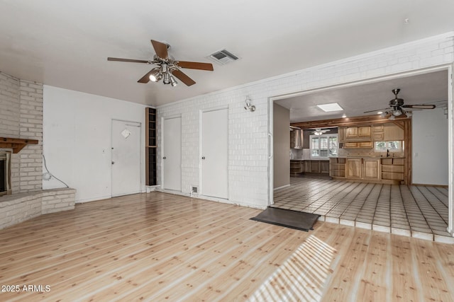 unfurnished living room with visible vents, brick wall, a brick fireplace, light wood-style flooring, and a ceiling fan