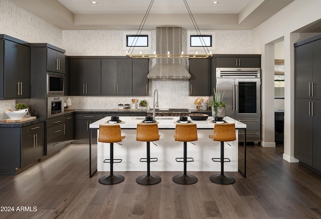 kitchen with a kitchen bar, wall chimney range hood, a center island with sink, built in appliances, and backsplash
