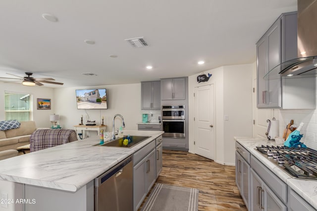 kitchen featuring gray cabinets, sink, a kitchen island with sink, stainless steel appliances, and wall chimney range hood