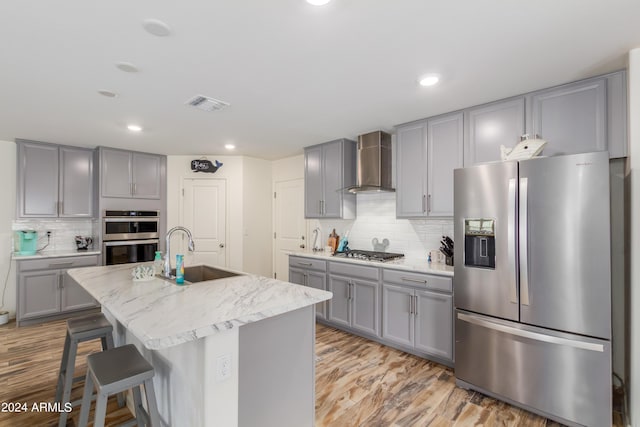 kitchen featuring sink, appliances with stainless steel finishes, a kitchen island with sink, light hardwood / wood-style floors, and wall chimney exhaust hood