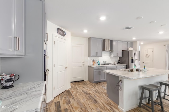 kitchen with a kitchen bar, gray cabinetry, a center island with sink, appliances with stainless steel finishes, and wall chimney range hood