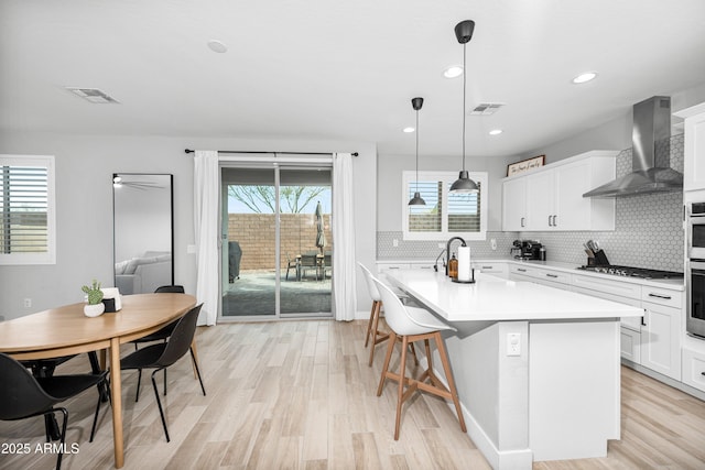 kitchen with pendant lighting, wall chimney range hood, a kitchen island with sink, white cabinetry, and stainless steel gas stovetop