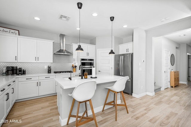 kitchen with wall chimney exhaust hood, decorative light fixtures, a center island with sink, stainless steel appliances, and white cabinets