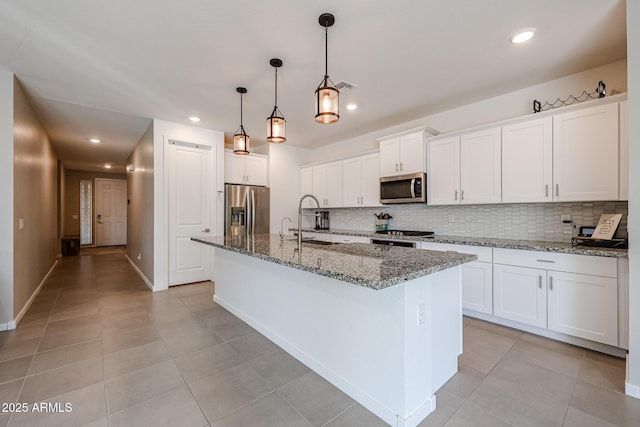 kitchen with a center island with sink, stainless steel appliances, white cabinetry, a sink, and dark stone counters