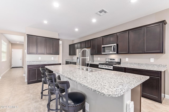 kitchen with appliances with stainless steel finishes, an island with sink, sink, light stone counters, and dark brown cabinets