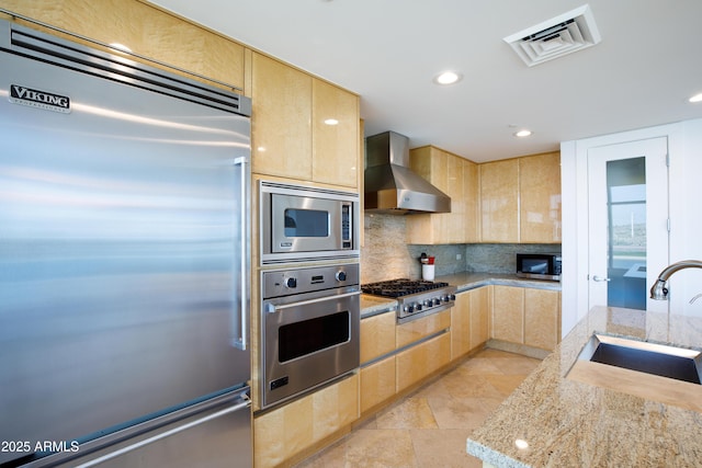 kitchen with sink, built in appliances, light stone countertops, and wall chimney range hood