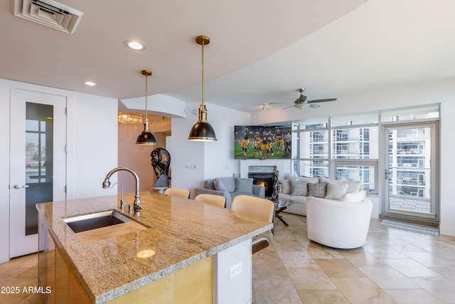 kitchen featuring light stone counters, ceiling fan, a kitchen island with sink, sink, and hanging light fixtures