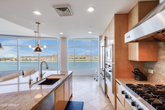 kitchen with light stone counters, wall chimney exhaust hood, sink, pendant lighting, and a water view