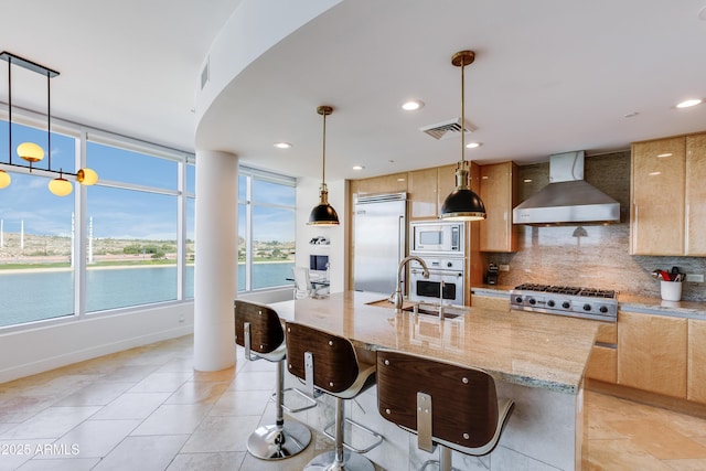 kitchen with sink, wall chimney range hood, built in appliances, decorative light fixtures, and a center island with sink