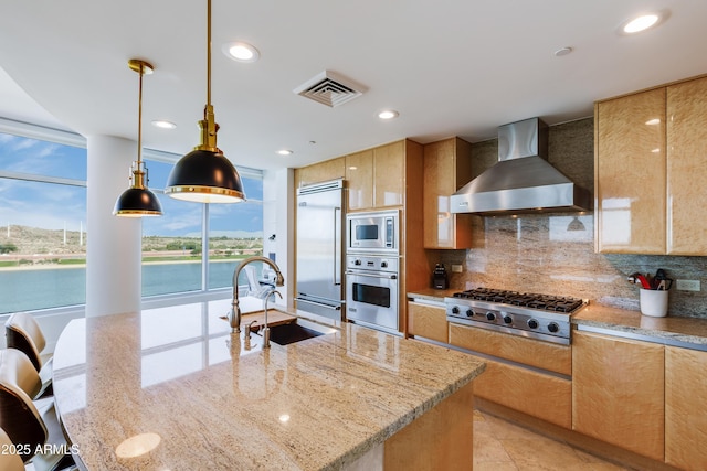 kitchen featuring pendant lighting, sink, built in appliances, wall chimney exhaust hood, and light stone counters