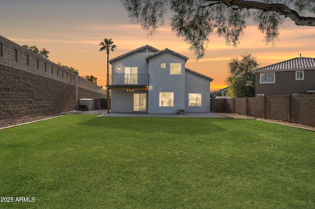 back of house at dusk with a fenced backyard, a lawn, a balcony, and stucco siding