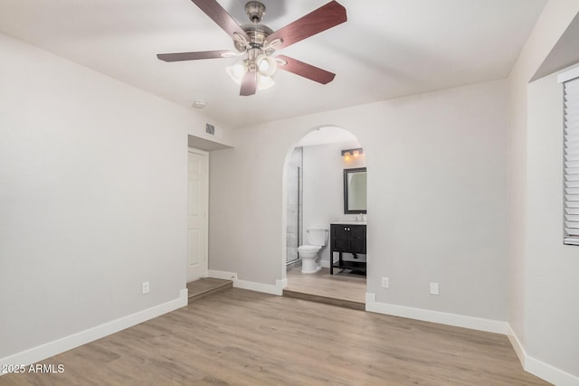 unfurnished bedroom with arched walkways, baseboards, light wood finished floors, ensuite bathroom, and ceiling fan