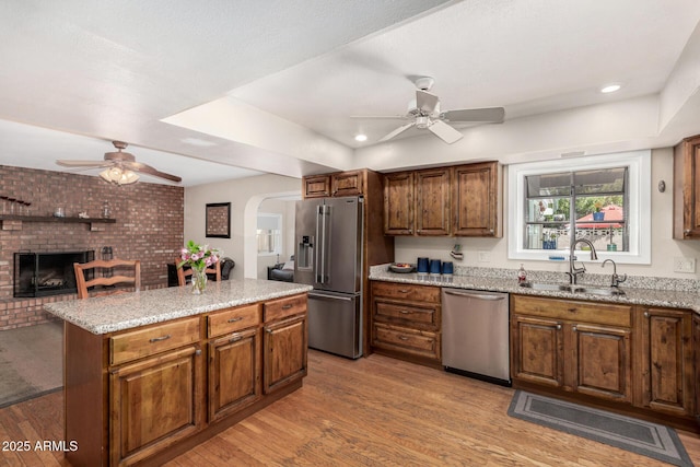 kitchen with a brick fireplace, light wood-type flooring, appliances with stainless steel finishes, arched walkways, and a sink