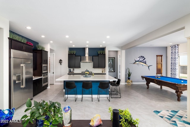 kitchen featuring wall chimney exhaust hood, a breakfast bar, tasteful backsplash, appliances with stainless steel finishes, and a kitchen island with sink