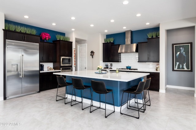 kitchen featuring wall chimney exhaust hood, a breakfast bar area, built in appliances, a center island with sink, and decorative backsplash