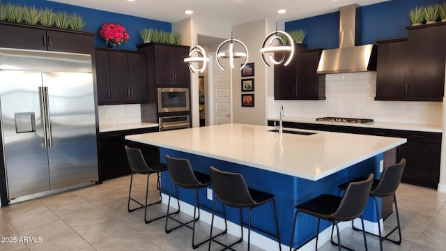 kitchen featuring wall chimney exhaust hood, sink, built in appliances, dark brown cabinets, and a kitchen island with sink