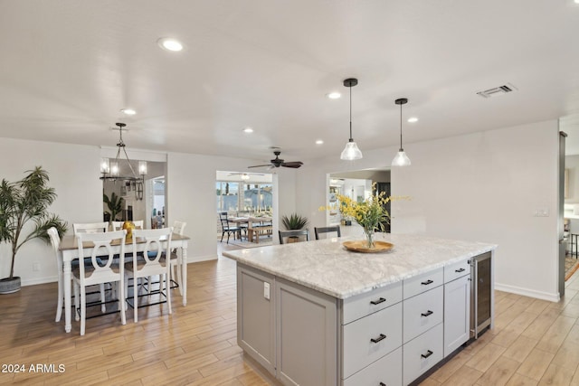 kitchen featuring wine cooler, light stone countertops, pendant lighting, ceiling fan with notable chandelier, and a center island