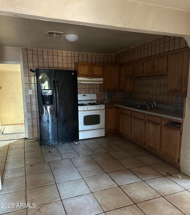 kitchen with tasteful backsplash, white gas range oven, black refrigerator with ice dispenser, under cabinet range hood, and a sink