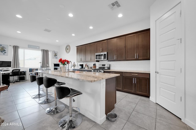 kitchen with a breakfast bar, a center island with sink, light tile patterned floors, appliances with stainless steel finishes, and light stone counters